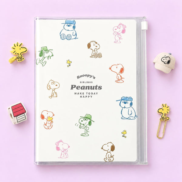 A Snoopy's Siblings Peanuts plastic organizer notebook with a clear cover, decorated with colorful cartoon character illustrations, displayed on a light pink background with themed props.