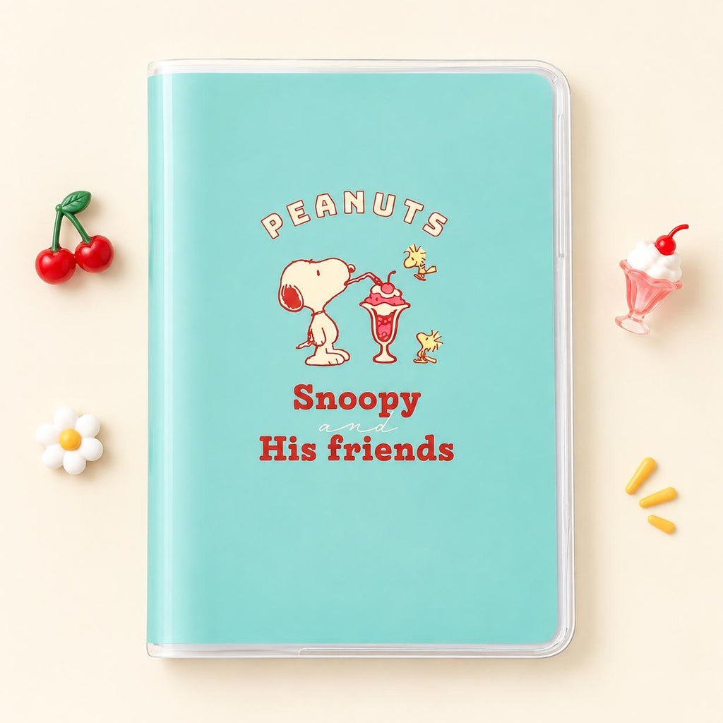 A mint green notebook with a clear plastic cover, featuring a retro Peanuts graphic of Snoopy and Woodstock with the text 'PEANUTS Snoopy His friends'. The notebook is styled in a flat-lay on a light beige background with decorative props including cherries, a daisy, and a milkshake.