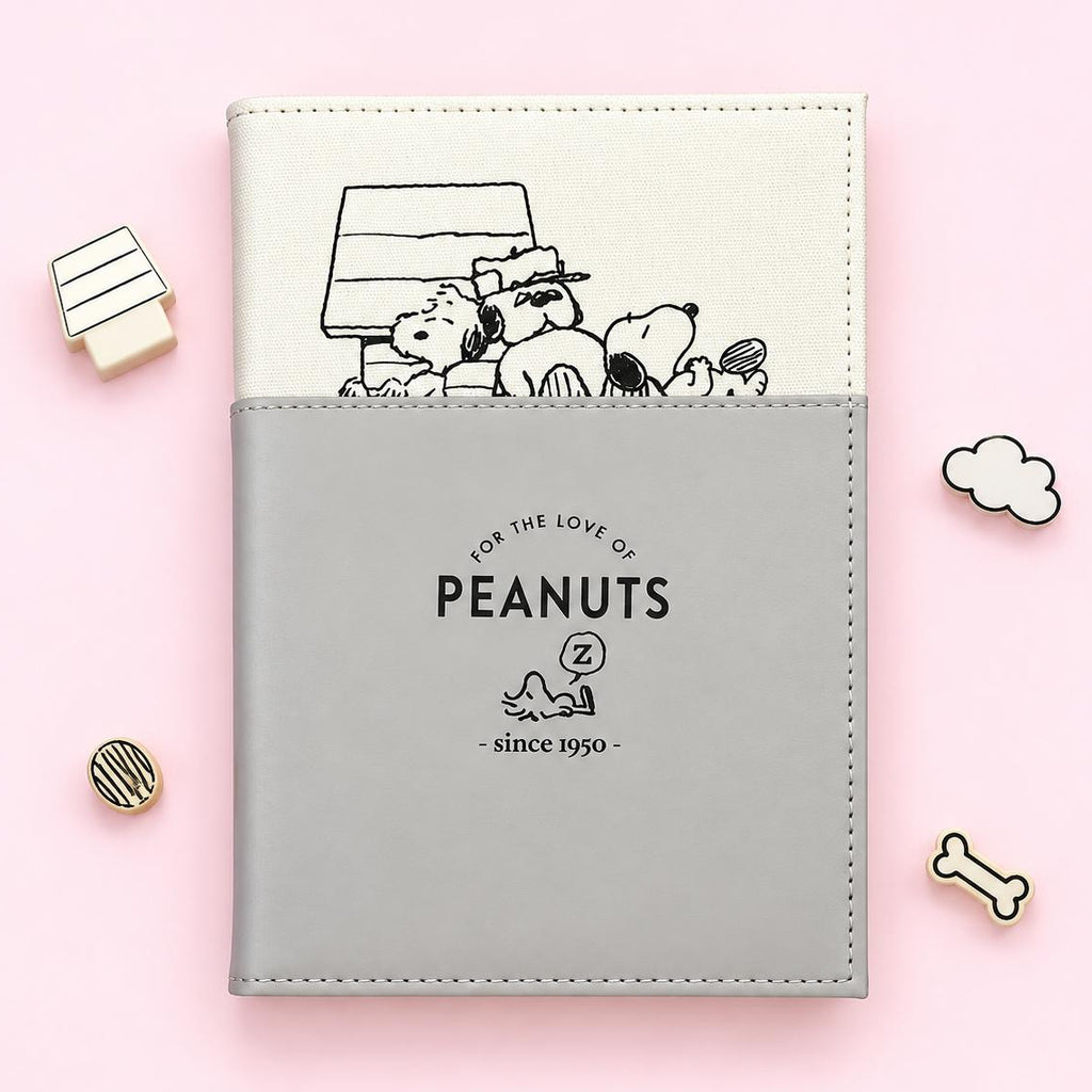 A Peanuts-themed notebook or planner cover on a light pink background, featuring a cream top section with a black line drawing of Peanuts characters and a gray bottom section with the text 'FOR THE LOVE OF PEANUTS' and 'since 1950'. Small decorative props are placed around it.