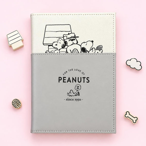 A Peanuts-themed notebook or planner cover on a light pink background, featuring a cream top section with a black line drawing of Peanuts characters and a gray bottom section with the text 'FOR THE LOVE OF PEANUTS' and 'since 1950'. Small decorative props are placed around it.