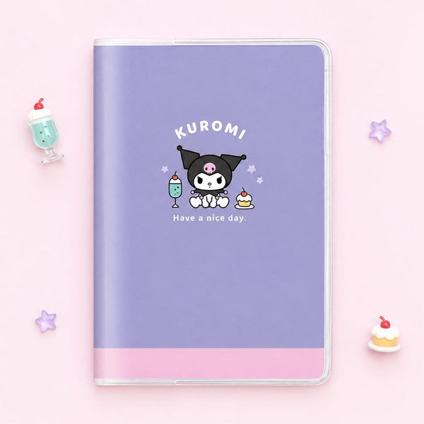 A pastel purple Sanrio schedule notebook with a pink band at the bottom and a Kuromi character graphic on the front, photographed from above on a light pink background with small decorative props around it.